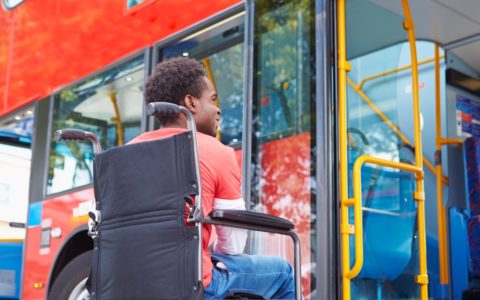 Fotografia digital colorida. Um homem negro, cabelos Black estiloso, usa calça jeans e blusa de manga comprida vermelha e branca. Ele esta sorrindo. Sentado em uma cadeira de rodas, aguarda no ponto de ônibus, a sua frente parte da lateral de um ônibus com detalhes amarelo.